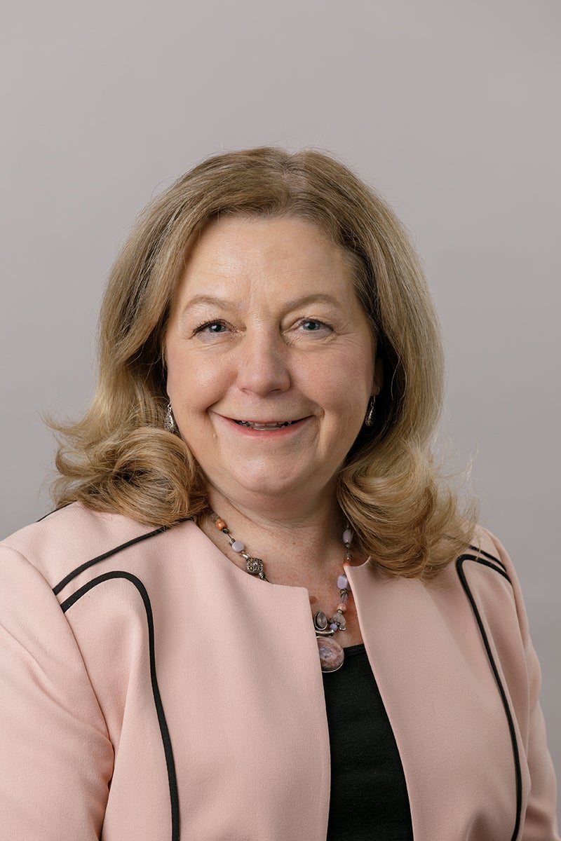 woman with shoulder-length light brown hair wearing a peach colored jacket with a black blouse underneath, smiling into the camera