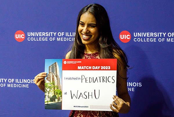 female student with brown hair holding a sign showing her residency match, in front of a backdrop with the College of Medicine logo