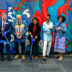 the named people standing in front of a colorful mural and using over-sized scissors to cut a large blue ribbon
