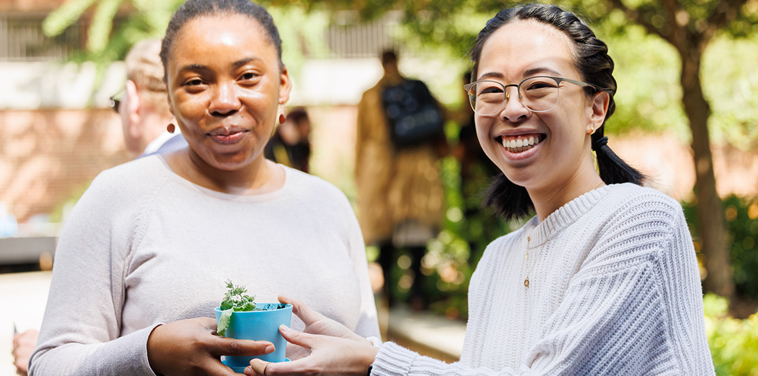 two women smiling and holding a small pot with a green plant in it