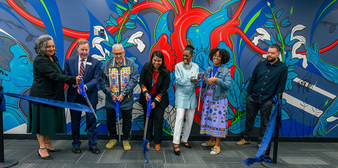 the named people standing in front of a colorful mural and using over-sized scissors to cut a large blue ribbon