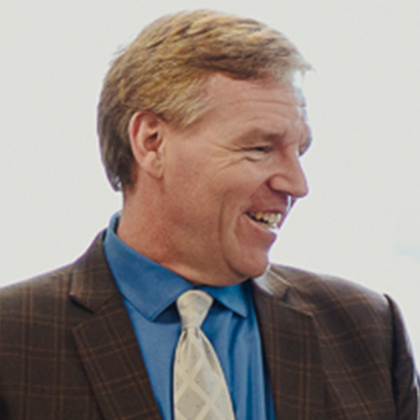 main with short sandy-colored hair smiling and wearing a brown plaid jacket, blue shirt and beige neck tie