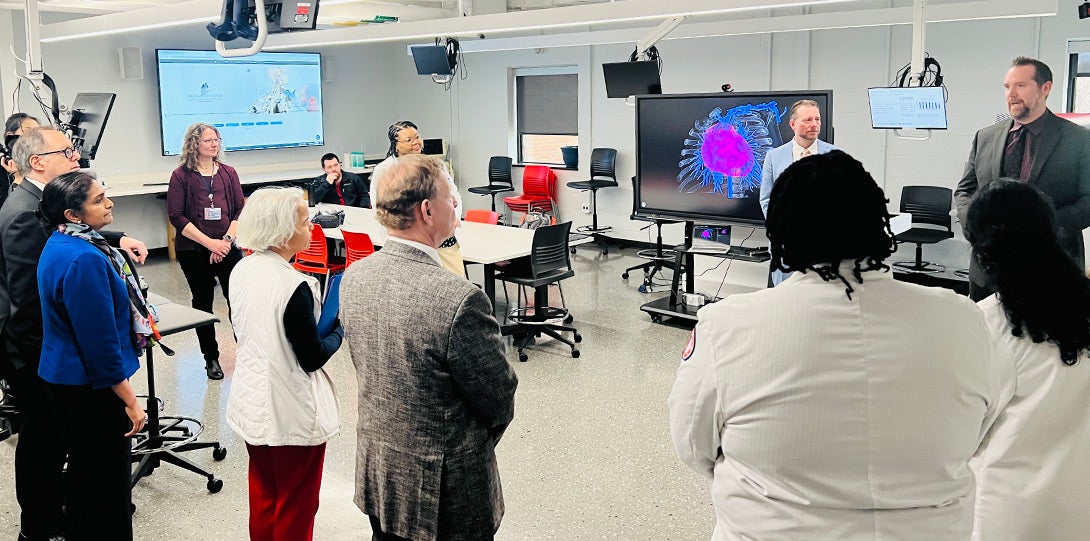 various people standing in a large classroom space, with a large video screen showing an electronic image of a human heart while a man in a dark gray suit explains