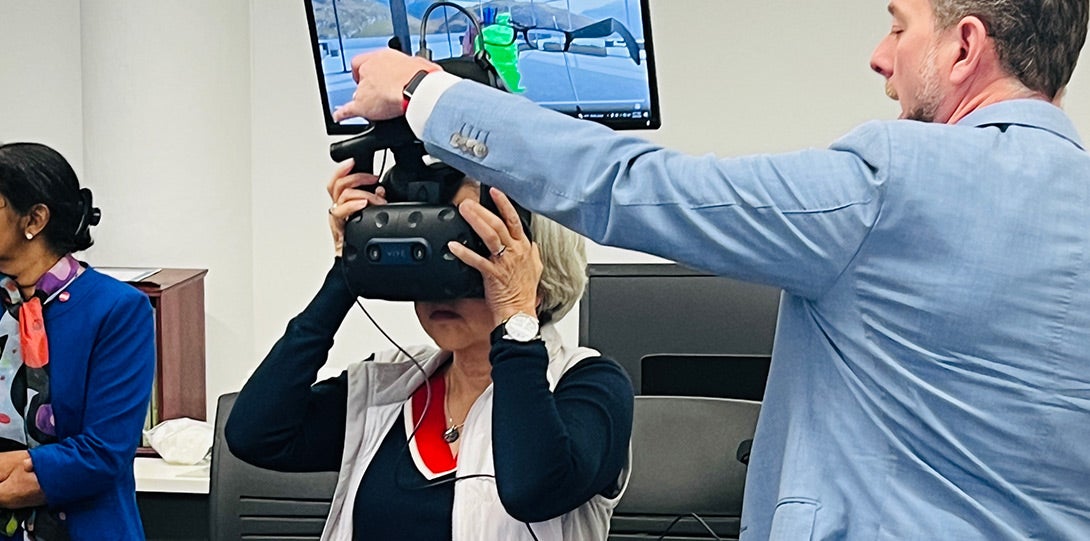 A man with short dark hair and light blue suit is fitting virtual reality goggles onto the head of a woman wearing a black blouse with a white jacket