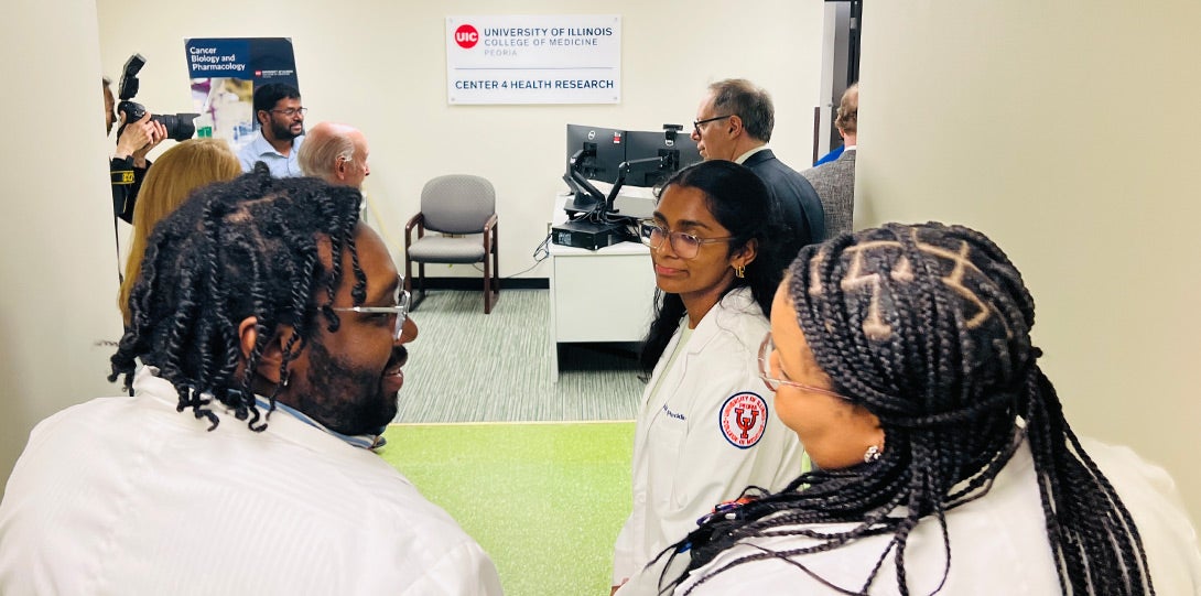 in the foreground, two African American medical students wearing white lab coat as they enter the center. In the background are other people in a modern office space.