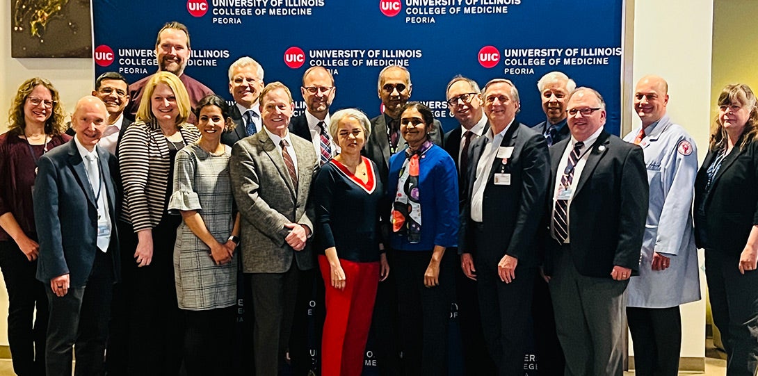 A large group of people in formal attire posing together in front of a background with repeating logos for the University of Illinois College of Medicine Peoria