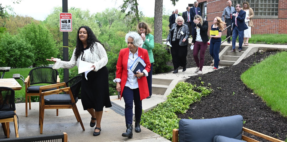 a young woman with long dark hair and wearing a black dress and white sweater is leading the chancellor, wearing a red jacket over a white blouse, and several other people through an outside courtyard