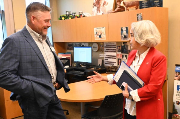 An office with a desk and computer in which a man with short dark hair, wearing a grey sport jacket, an oxford shirt and jeans, speaking with a woman who has shoulder-length white hair and wearing a white blouse under a red jacket. In the background is a sign saying RMED”