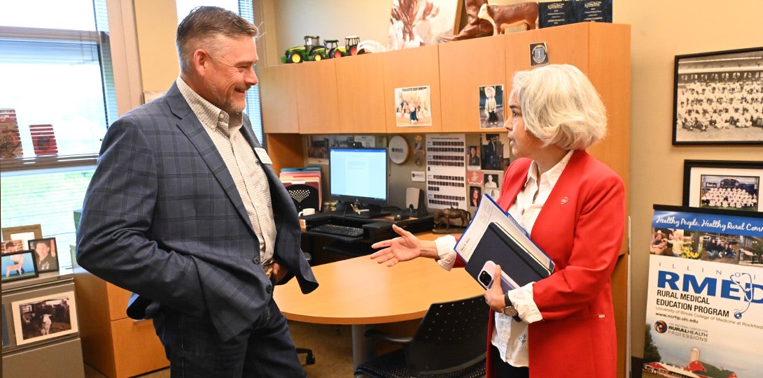 A mean wearing a blue checked sport coat and oxford shirt standing wiht his hands in his pockets, listening to the chancellor with short white hair and a red jacket with a white blouse. They are standing together in an office with a desk and computer.