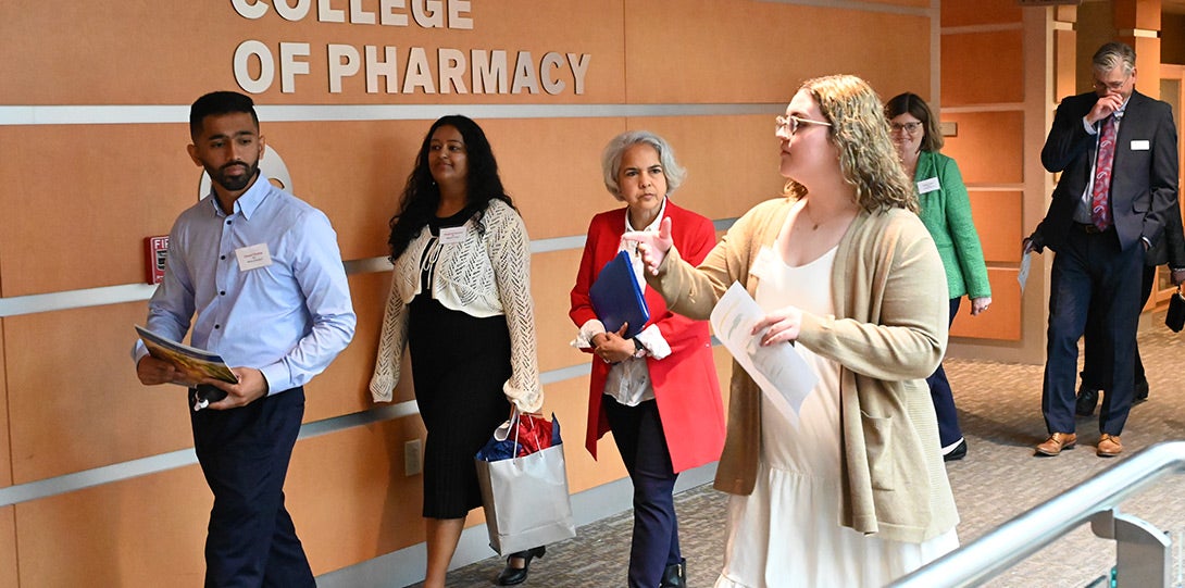 A young man with dark hair and a beard and wearing a blue shirt, a woman with long dark hair and wearing a white sweater over a black dress, a Latinx woman with white hair and weapon a red jacket over a white blouse, and a woman with long sandy-colored hair wearing a brown sweater, all walking together in a wood paneled hallway with a sign reading 