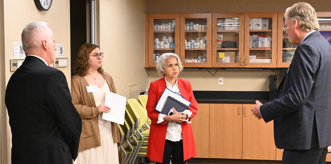 A man with short white hair and wearing a dark suit, a young woman with long brown hair and wearing a tan sweater over a white dree, a woman with shoulder length white hair wearing a red jacket over a white blouse, and man with salt and pepper hair hearing a dark gray suit, all standing in a laboratory space with shelves in the background filled with pharmaceutical bottles.