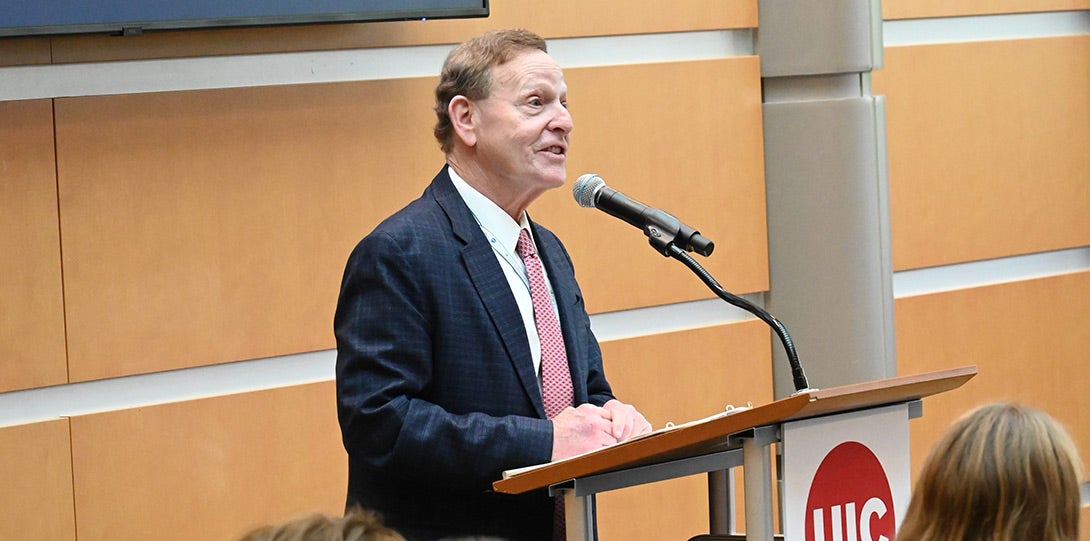 a man with short brown hair and wearing a blue suit coat, white shirt and red neck tie, standing behind a podium and speaking into a microphone