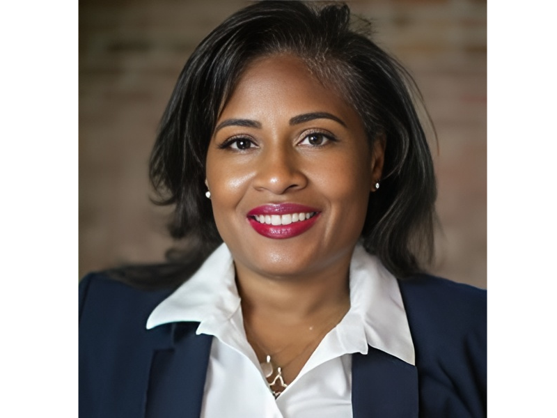 African American woman with shoulder length black hair and wearing a blue suit jacket and white blouse, posing in front of a brick wall and smiling into the camera