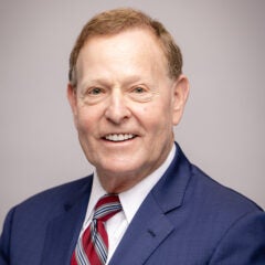 a man with short brown hair wearing a blue suit jacket, white shirt and blue and red neck tie, smiling into the camera