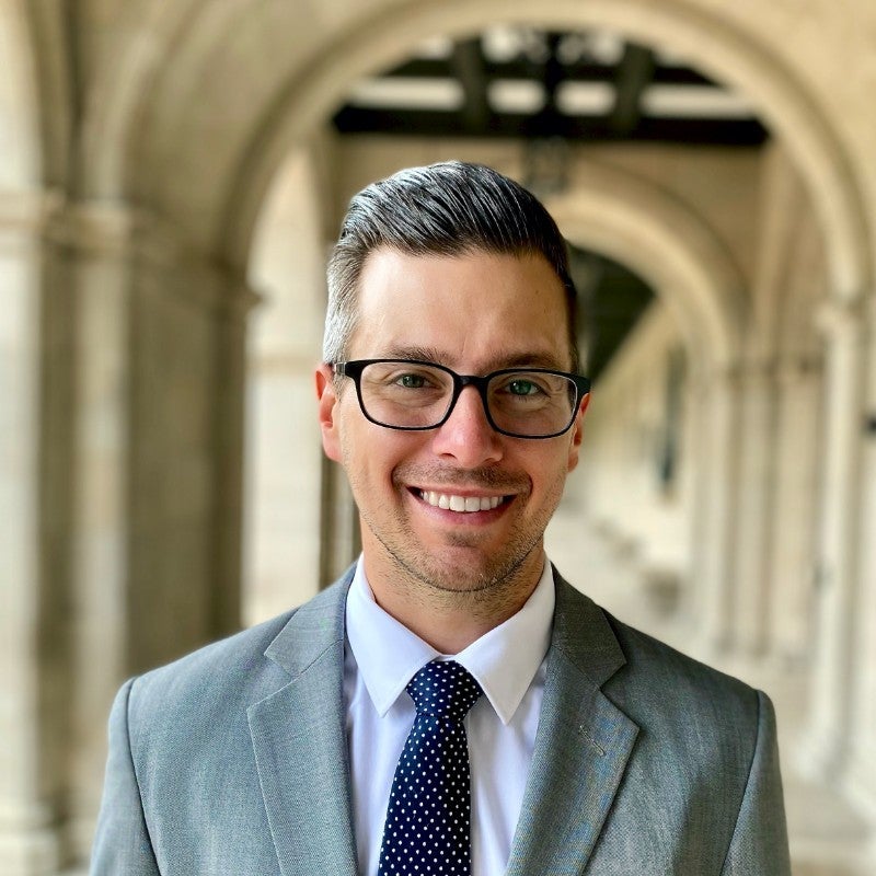 younger man with short dark hair and glasses, wearing a light gray suit, white shirt and bliue necktie, standing in an outdoor academic corridor