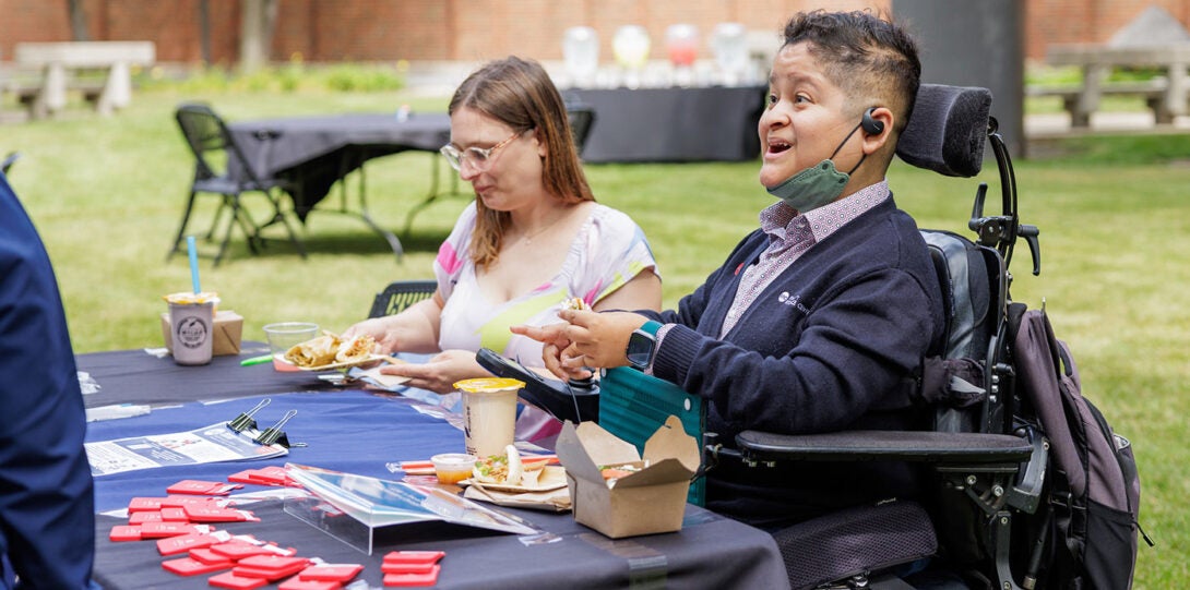 A young man in a power wheelchair, sitting behind a table covered with information and giveaway, smiling and speaking with an attendee