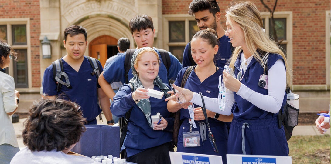 A diverse group of students in dark blue scrubs, standing at an exhibitor table and taking giveaways