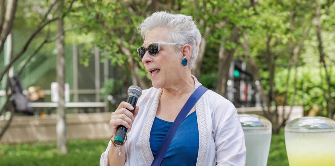 A woman with white hair, wearing sunglasses and a blue blouse with white sweater, speaking into a microphone