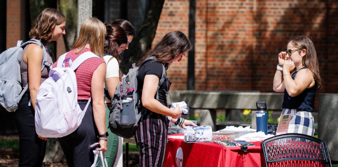 a group of young women looking at information and giveaways on a table adorned with a red table cloth