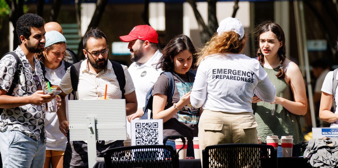 A diverse group of young poeple standin at a table, with a woman behind the table wearing a tee shirt that says Emergency Preparedness