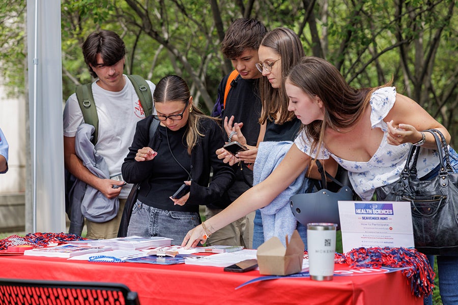 A diverse group of young students standing around a table adorned with a red table cloth and various items of information and giveaways