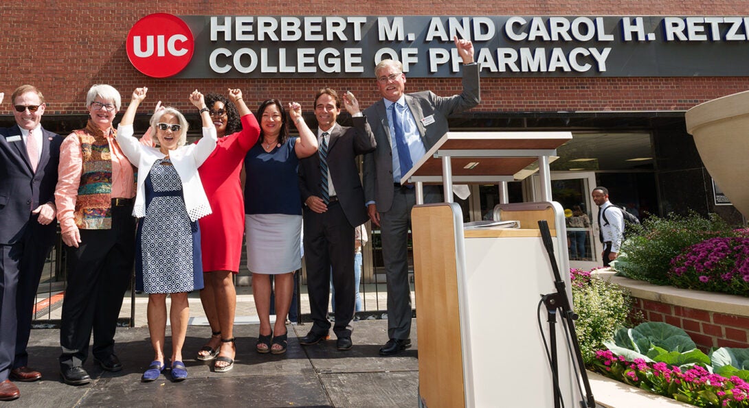 A group of seven women and men standing on a stage with their arms upraised in a cheer. Behind them is the new illuminated sign reading UIC Herbert M. and Carol H. Retzky College of Pharmacy.