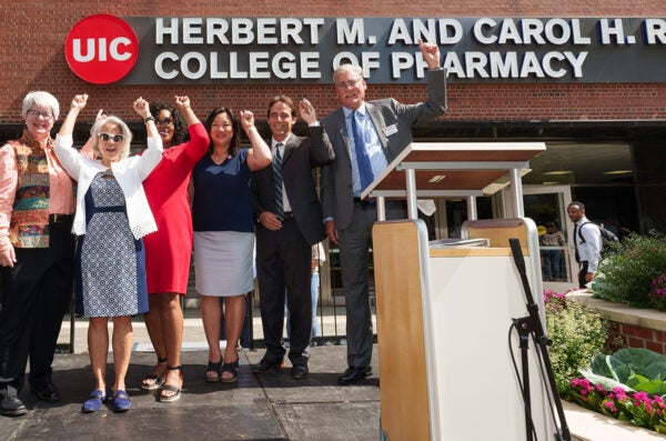 A group of seven women and men standing on a stage with their arms upraised in a cheer. Behind them is the new illuminated sign reading UIC Herbert M. and Carol H. Retzky College of Pharmacy.