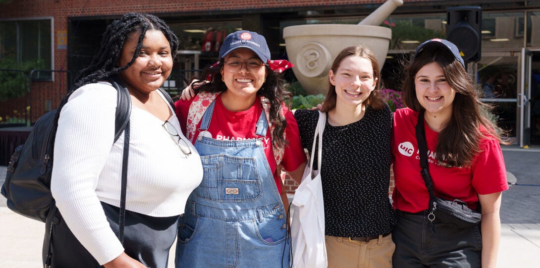 Diverse group of four young women standing in front of the College of Pharmacy. Two are wearing College of Pharmcy ball caps. Behind them is a large concrete sculpture of a mortar and pestle.