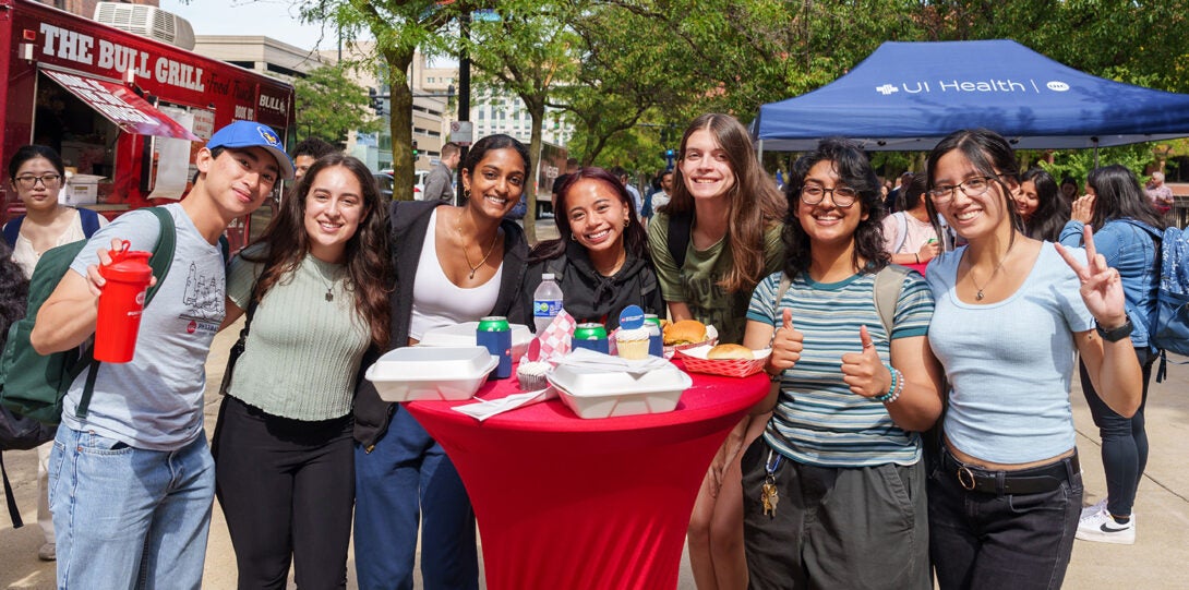 A diverse group of students with food and cans of soda, posing a smiling around a highboy table covered with with red table cloth.