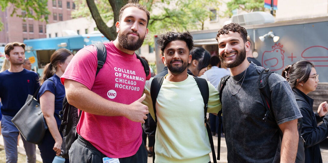 Three male students, one wearing a College of Pharmacy Rockford t-shirt, smiling and posing together in front of food trucks