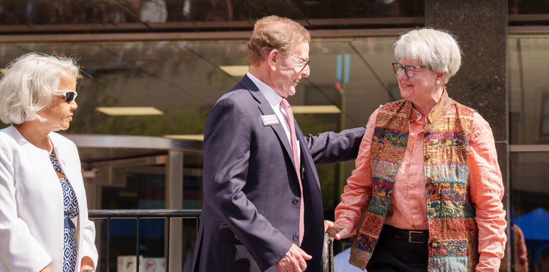 At right a woman in a white jacket; at center, a man in a dark gray suite; at right, a woman wearing a salmon colored blouse and colorful vest.