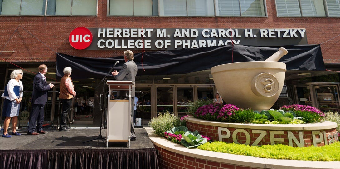 Four people on a stage in front of a brick building. On the front of the building, a black drape is falling to reveal a sign which reads: UIC Herbert M. and Carol H. Retzky College of Pharmacy.