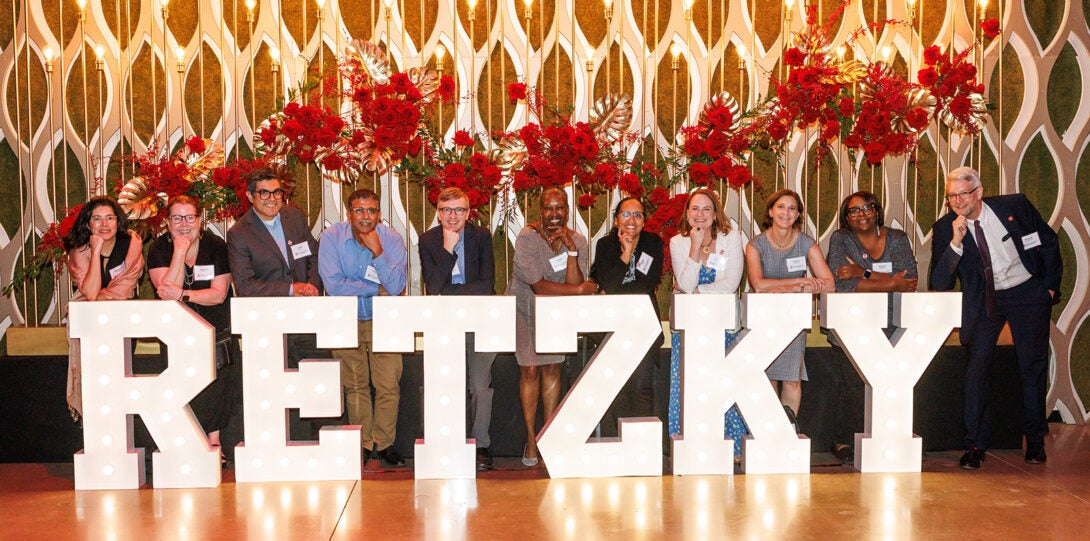 Eleven people in formal attire, all standing behind a set of large white letters spelling Retzky. Behind them is a patterned wall adorned with red flowers.