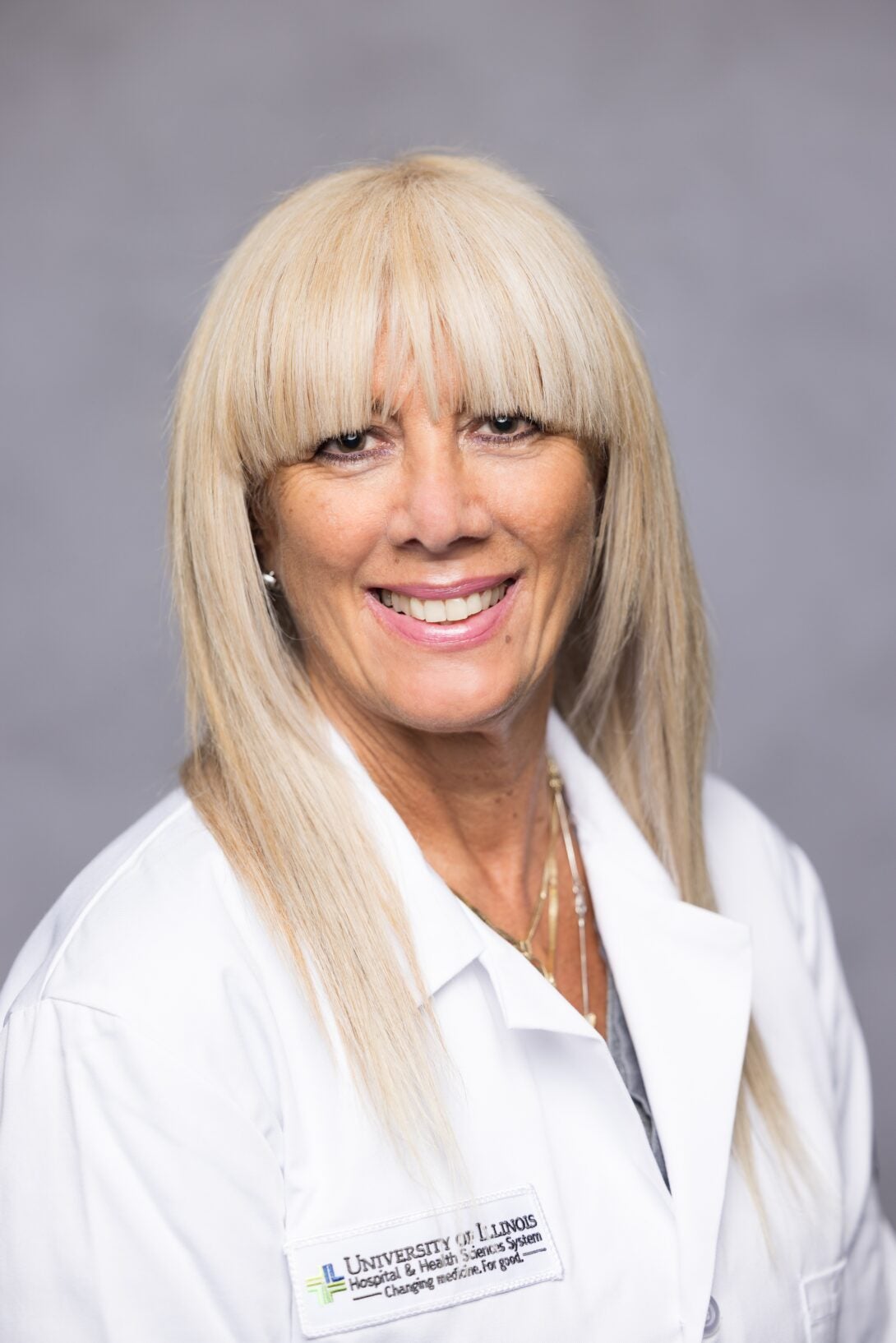 Doctor with long blonde hair and bangs smiles at the camera while wearing a white lab coat.