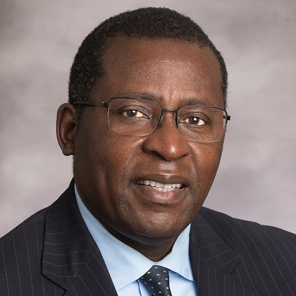 African American man with short dark hair wearing a pair of glasses, a gray suit jacket, light blue shirt and blue neck tie